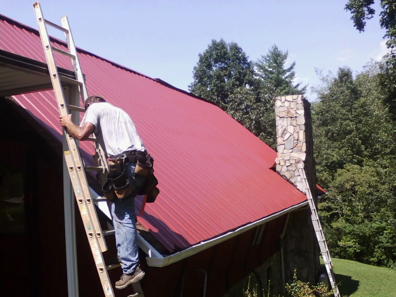 Roofer on a ladder beside a red metal roof and stone chimney — Soffit & Fascia Repair by Roofing Brothers in Dutton